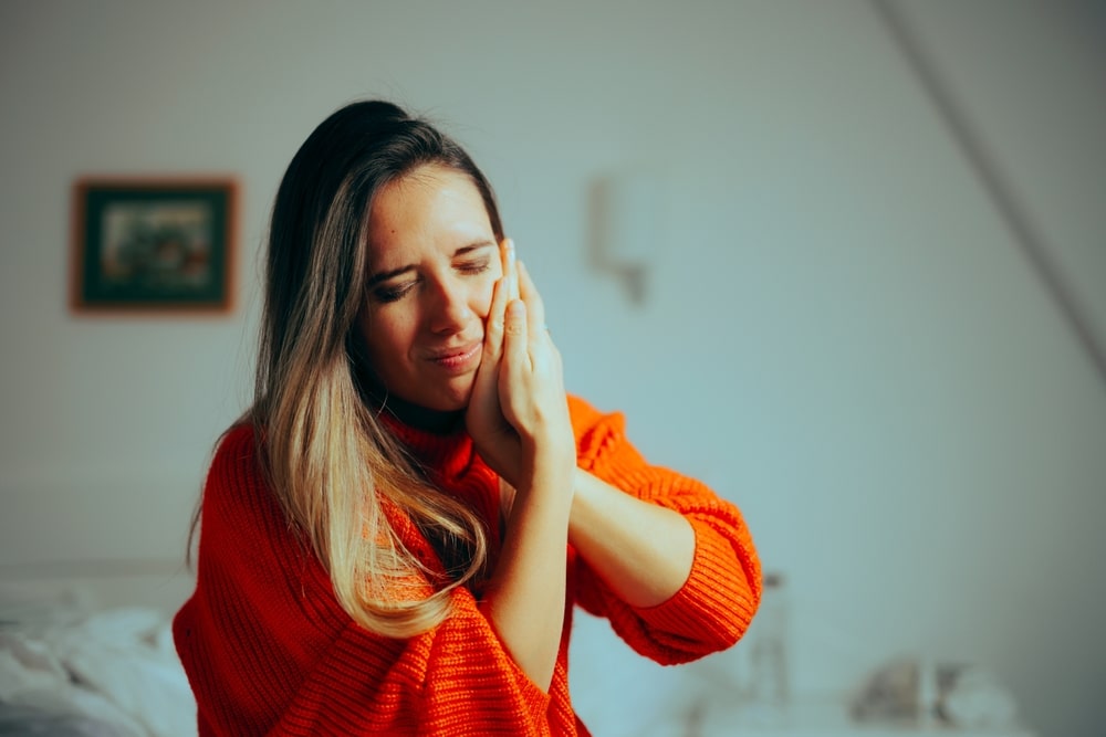 Young woman wincing and clutching her jaw in pain from TMJ disorder symptoms, in need of relief.